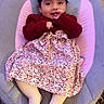 child, baby, toddler, portrait, sitting, baby_seat, smiling, face, red_bow, dark_hair, sweater, floral_dress, striped_tights, pink_cushion, indoor, wooden_floor, rug, furniture, finger_in_mouth, cozy