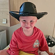 Carter joined the competition — help win amazing prizes! child, boy, cowboy_hat, red_shirt, yellow_shorts, smile, indoor, bed, cross_legged, radiator, cabinet, furniture, toy, plush_toy, casual_clothing, light_skin, short_hair, happy, portrait, home