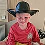 child, boy, cowboy_hat, red_shirt, yellow_shorts, smile, indoor, bed, cross_legged, radiator, cabinet, furniture, toy, plush_toy, casual_clothing, light_skin, short_hair, happy, portrait, home