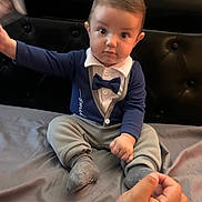 Edi joined the competition — help win amazing prizes! baby, child, infant, bow_tie, blue_clothing, sitting, bed, hand, curious, indoor, portrait, person, headboard, gray_pants, sock, furniture, young_child, cute, engagement, focus
