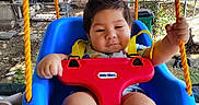 Jose is registered to the contest to win money with this photo: baby, child, infant, swing, outdoor, backyard, smiling, seated, playground, blue_swing, red_safety_bar, yellow_ropes, harness, portrait, face, hands, legs, trees, fence, sunlight