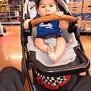 Jose is registered to the contest to win money with this photo: aisle, baby, bare_feet, blue_shirt, boxes, cap, child, face, indoors, infant, paper_towels, portrait, retail, safety_harness, shopping, sitting, smiling, store, stroller, stroller_bar