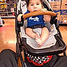 Jose is registered to the contest to win money with this photo: baby, child, stroller, cap, blue_shirt, face, portrait, sitting, indoors, store, shopping, aisle, paper_towels, boxes, safety_harness, stroller_bar, bare_feet, infant, retail, smiling