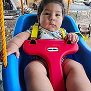Jose is registered to the contest to win money with this photo: baby, child, infant, swing, playground, outdoor, sitting, safety_harness, red_bar, blue_seat, serious_expression, dark_hair, portrait, close_up, backyard, fence, trailer, play_equipment, summer, photography