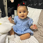 Tanisha is registered to the contest to win money with this photo: baby, bed, blue_dress, child, comfortable, cozy, cute, face, hair_bow, happy, indoors, infant, person, pillow, portrait, sitting, smiling, soft_lighting, white_socks, young_child