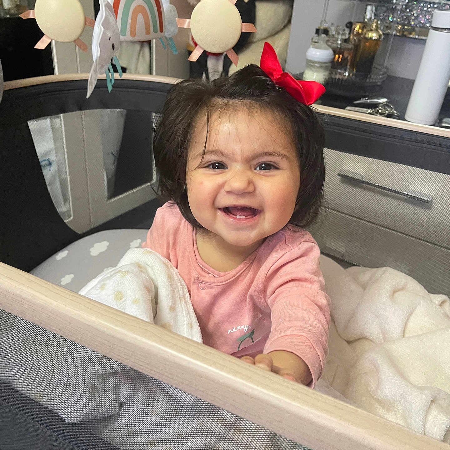 Tanisha is registered to the contest to win money with this photo: baby, blanket, bottle, child, cozy, face, furniture, hair, happy, indoor, mirror, person, pink_clothing, playpen, red_bow, smile, table, teddy_bear, toy_mobile