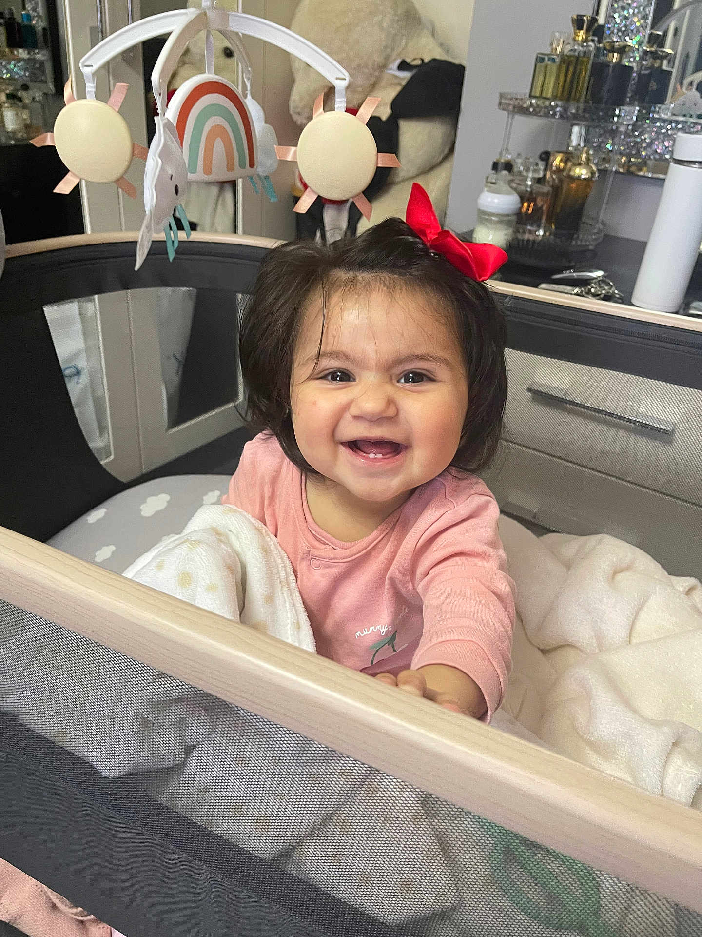 Tanisha is registered to the contest to win money with this photo: baby, blanket, bottle, child, cozy, face, furniture, hair, happy, indoor, mirror, person, pink_clothing, playpen, red_bow, smile, table, teddy_bear, toy_mobile