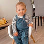 toddler, child, indoor, chair, denim_overalls, striped_shirt, sneakers, white_shoes, wood_floor, table, dining_chair, clock, plant, modern_furniture, cute, smiling, person, young_child, casual_clothing, home