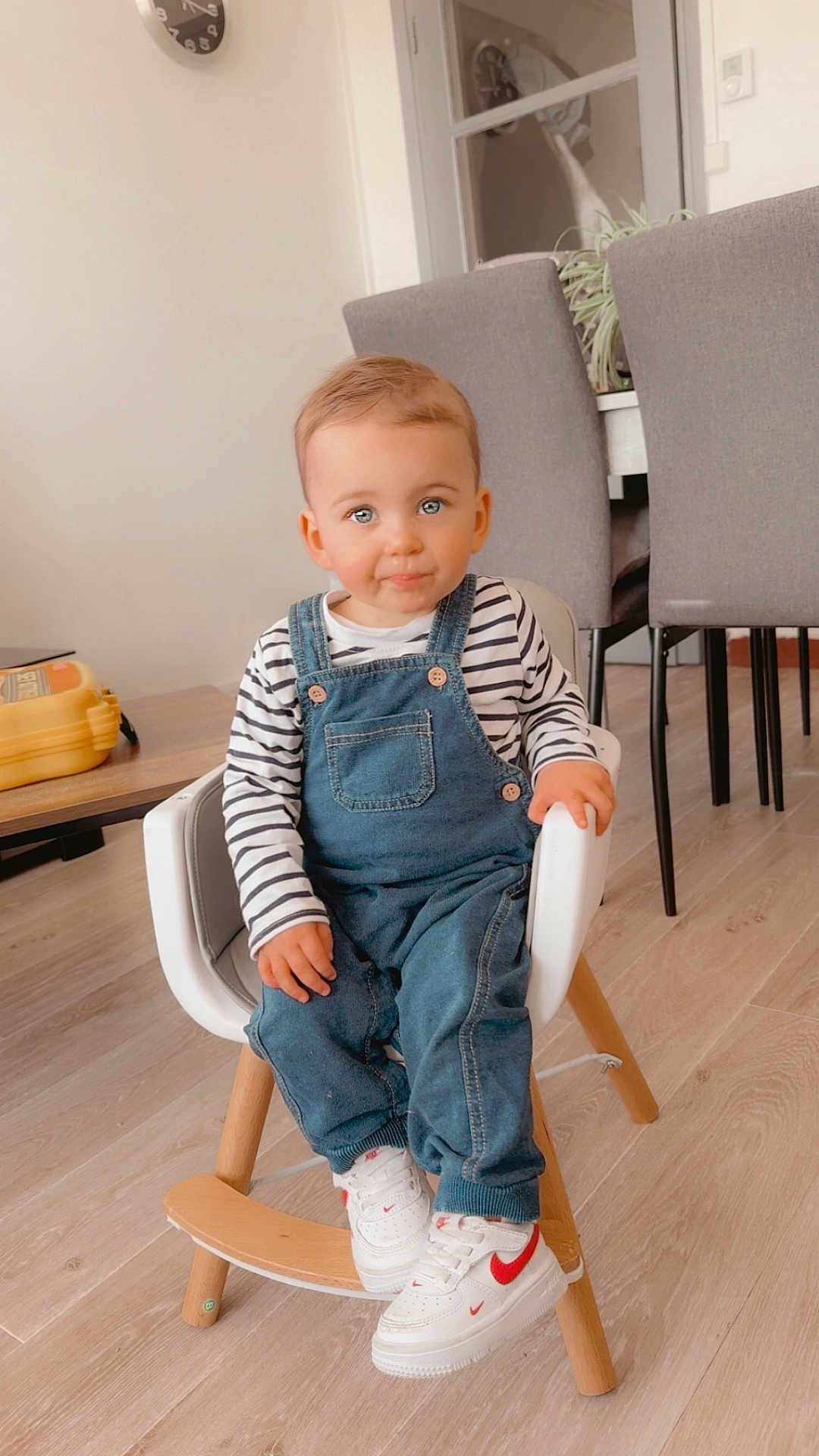 Kassïm a rejoint le concours — aidez-le/la à gagner de superbes lots ! toddler, child, indoor, chair, denim_overalls, striped_shirt, sneakers, white_shoes, wood_floor, table, dining_chair, clock, plant, modern_furniture, cute, smiling, person, young_child, casual_clothing, home