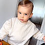 toddler, child, blue_eyes, sweater, indoor, wooden_table, pen, curious, hair, face, person, furniture, chair, light, window, curtain, skin, expression, casual, young
