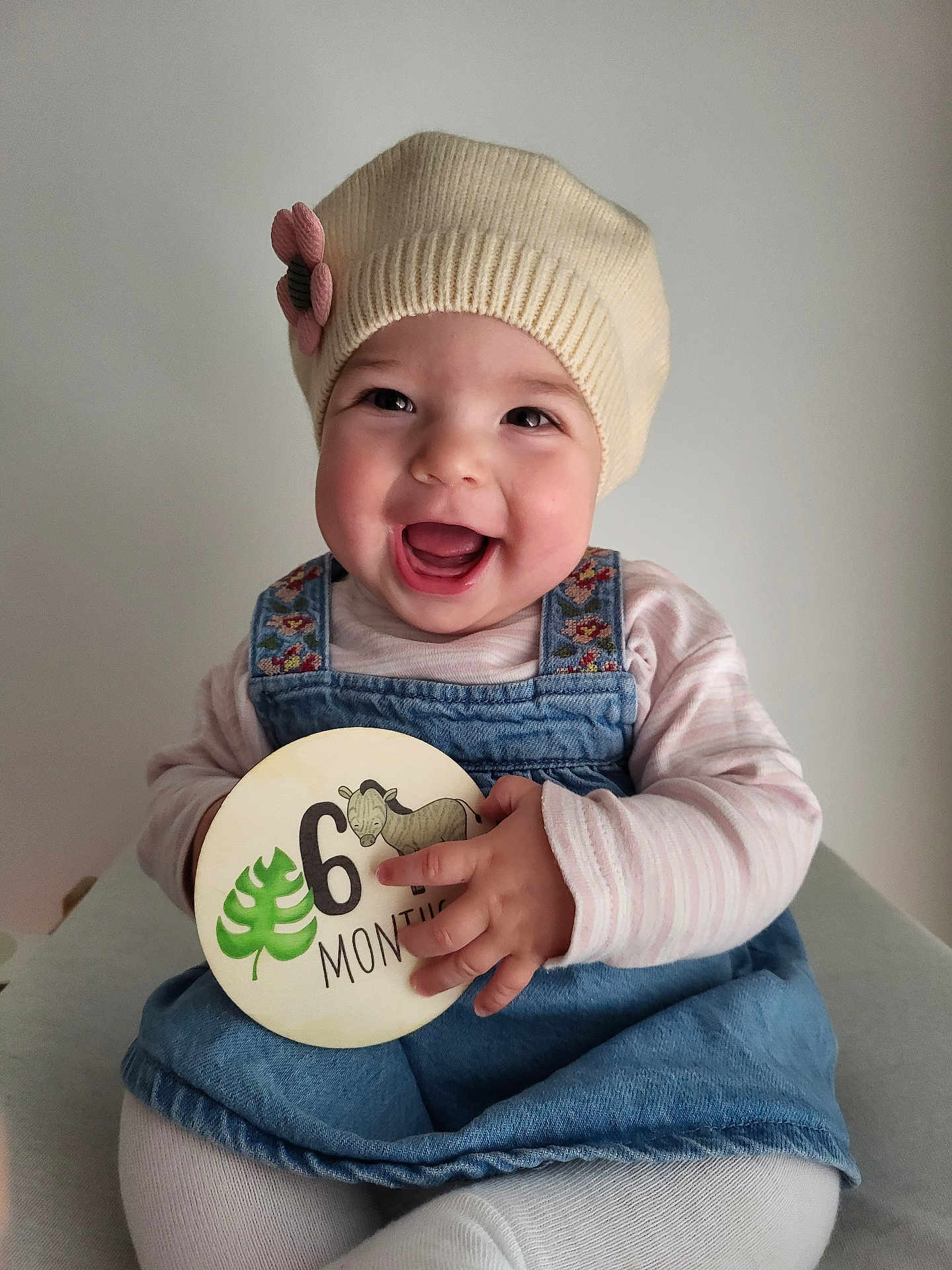Victoria participe au concours pour gagner de l'argent avec cette photo : baby, child, smiling, happy, hat, knit_hat, flower, denim_overalls, clothing, round_sign, wooden_sign, months, hand, face, portrait, sitting, indoor, cute, infant, person