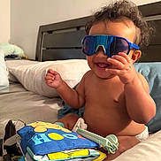 Zayn is registered to the contest to win money with this photo: baby, child, infant, sunglasses, smiling, laughing, sitting, bed, bedroom, pillow, blanket, toy, cloth_book, colorful, portrait, indoor, home, bare_chested, diaper, playful