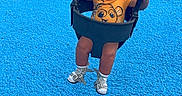 Zayn is registered to the contest to win money with this photo: child, toddler, playground, swing, smiling, sitting, outdoor, play_equipment, swing_chains, bucket_swing, orange_shirt, cartoon_shirt, sandals, socks, curly_hair, blue_surface, red_surface, playground_mat, playground_bench, daylight