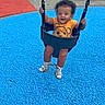child, toddler, playground, swing, smiling, sitting, outdoor, play_equipment, swing_chains, bucket_swing, orange_shirt, cartoon_shirt, sandals, socks, curly_hair, blue_surface, red_surface, playground_mat, playground_bench, daylight