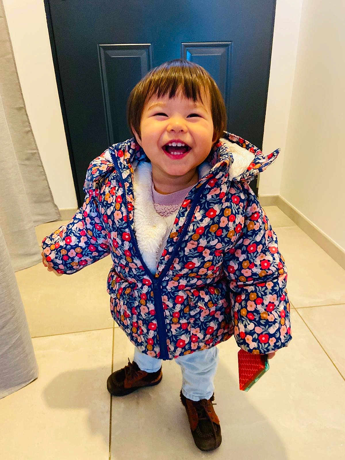 Luna participe au concours pour gagner de l'argent avec cette photo : toddler, child, smiling, jacket, floral_pattern, indoor, door, tile_floor, happy, holding_object, brown_shoes, pants, cute, standing, warm_clothing, short_hair, playful, young, person, portrait