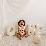 Amaya joined the competition — help win amazing prizes! baby_clothes, balloon, celebration, child, curly_hair, cute, decor, fabric, floor, happy, indoor, letters, minimalist, one, portrait, sitting, smiling, toddler, white_wall, wooden_floor