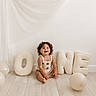 baby_clothes, balloon, celebration, child, curly_hair, cute, decor, fabric, floor, happy, indoor, letters, minimalist, one, portrait, sitting, smiling, toddler, white_wall, wooden_floor