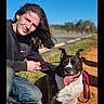 person, dog, leash, outdoor, sunny, grass, road, blue_sky, casual_clothing, happy, tongue_out, sitting, nature, smiling, young_adult, black_and_white_dog, jeans, nike_sweatshirt, sidewalk, daylight