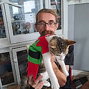 Marie is registered to the contest to win money with this photo: man, cat, glasses, mustache, indoor, cabinet, collectibles, clothing, table, box, furniture, pet, costume, red, green, white, brown, holding, sitting, person