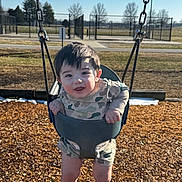 Karter is registered to the contest to win money with this photo: child, baby, swing, swing_set, playground, park, smile, happy, outdoor, wood_chips, chains, swing_seat, camouflage_clothing, infant, young_child, sunlight, grass, fence, face, portrait