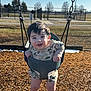 child, baby, swing, swing_set, playground, park, smile, happy, outdoor, wood_chips, chains, swing_seat, camouflage_clothing, infant, young_child, sunlight, grass, fence, face, portrait