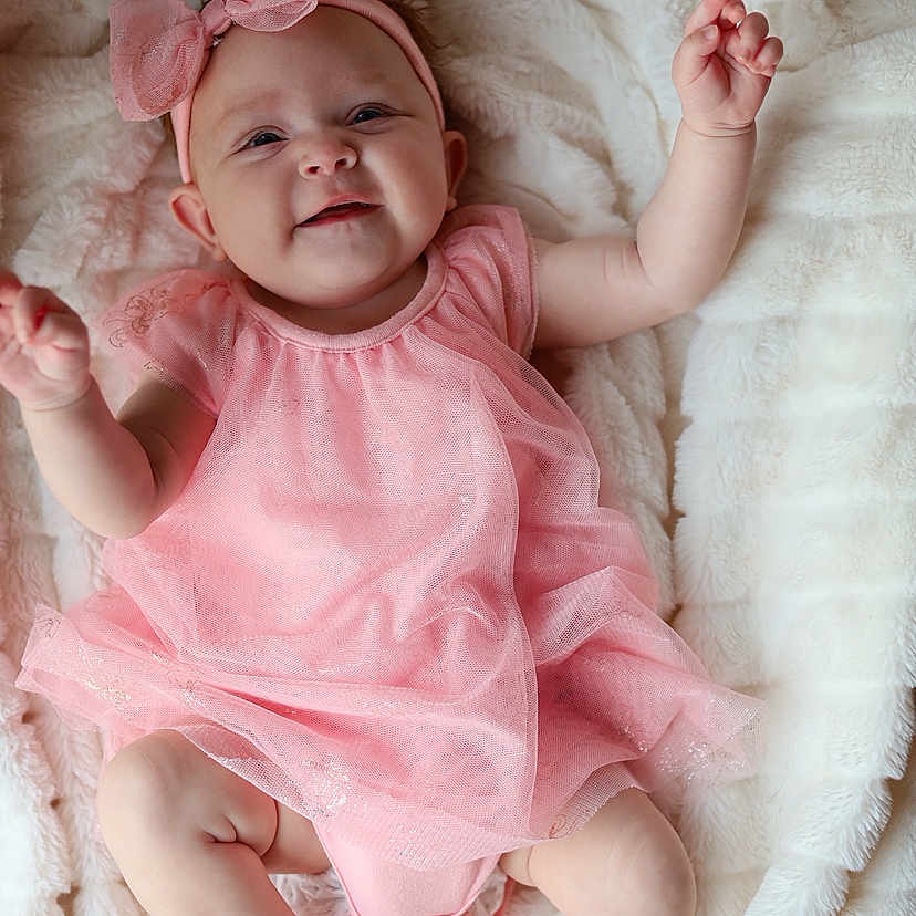 Milani is registered to the contest to win money with this photo: adorable, baby, blanket, bow, child, cozy, cute, feet, happy, headband, indoors, infant, lying_down, pink_dress, portrait, skin, small_hands, smiling, soft_texture, toddler