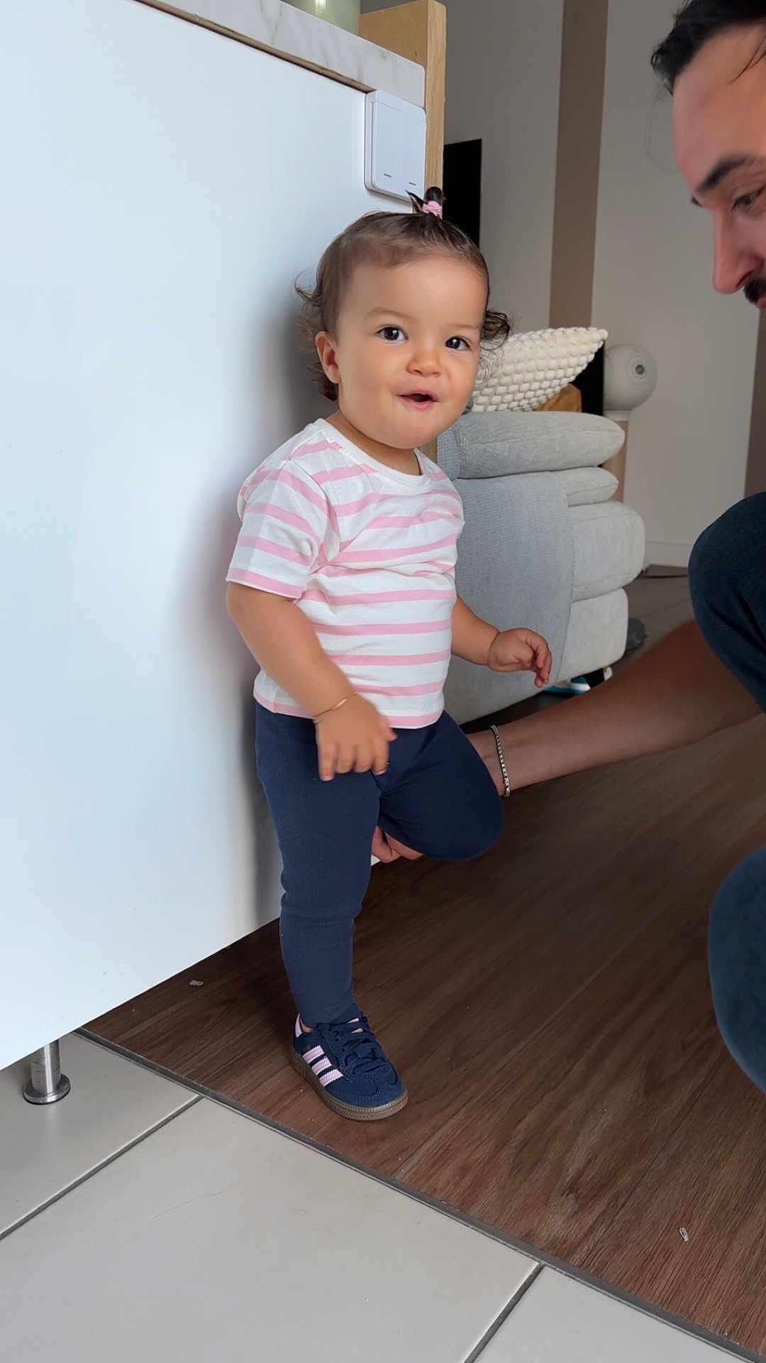Camélia a rejoint le concours — aidez-le/la à gagner de superbes lots ! toddler, child, indoor, person, standing, striped_shirt, navy_pants, sneakers, pony_tail, floor, adult, support, smile, happy, wood_floor, tile_floor, bracelet, couch, home, light