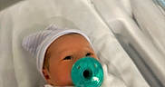 Isabella is registered to the contest to win money with this photo: baby, infant, newborn, pacifier, swaddled, blanket, hat, striped_hat, sleeping, face, eyes, portrait, hospital, bassinet, bed, indoors, soft_lighting, turquoise, white_clothing, close_up