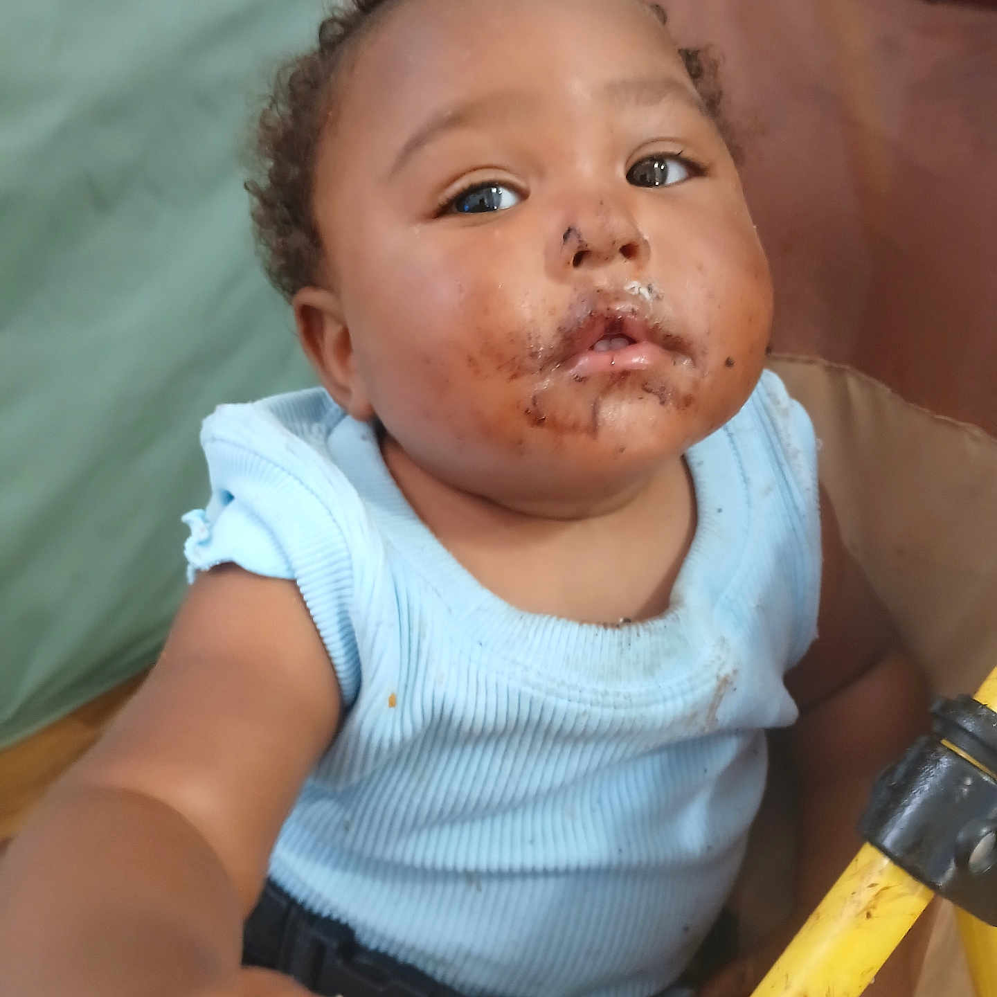 Dior is registered to the contest to win money with this photo: baby, child, infant, portrait, sitting, chair, face, curly_hair, smiling, curious, blue_clothing, sleeveless_top, food, crumbs, milk_mustache, snack, toy, yellow_object, indoor, home