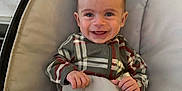 Huxley is registered to the contest to win money with this photo: baby, child, smiling, plaid, onesie, cushion, baby_seat, happy, infant, person, indoor, cute, clothing, small_hands, young, portrait, sitting, face, joyful, cozy