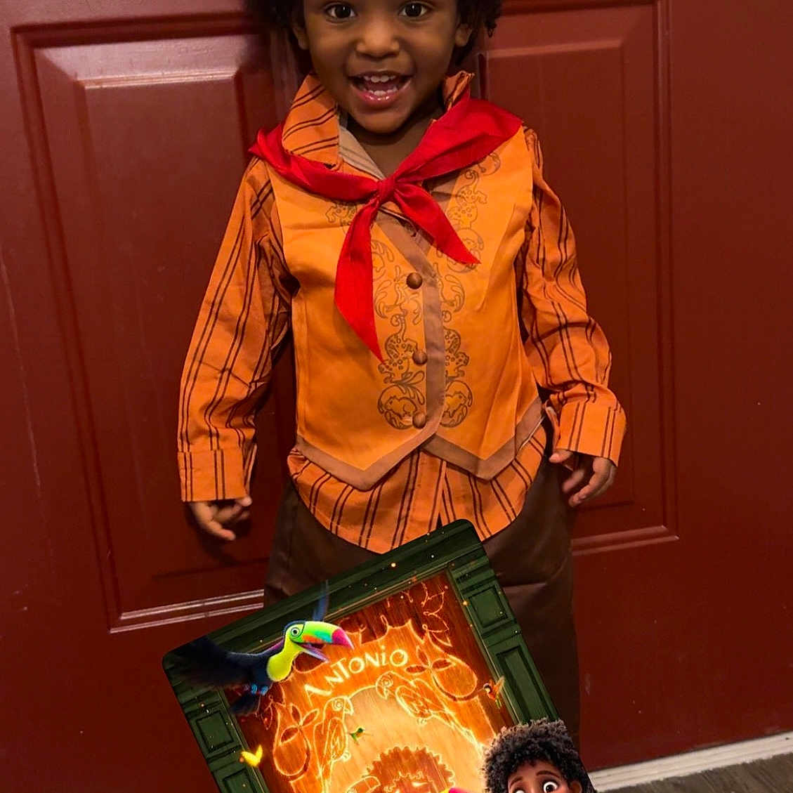 Cam is registered to the contest to win money with this photo: child, smiling, costume, orange_shirt, red_scarf, curly_hair, door, wood_floor, movie_poster, encanto, disney, toucan, animated_character, indoor, portrait, happy, boy, standing, colorful, playful