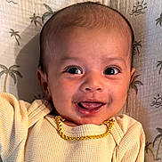 Amiri is registered to the contest to win money with this photo: baby, smile, face, eyes, gold_chain, cream_outfit, infant, lying_down, blanket, pattern, palm_tree, happy, cute, child, portrait, indoors, closeup, person, young, cheerful