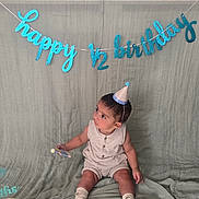 Faris joined the competition — help win amazing prizes! baby, child, party_hat, birthday, banner, celebration, half_birthday, sitting, cute, indoors, textile, background, decor, sock, small_object, portrait, infant, happy, one_half, milestone