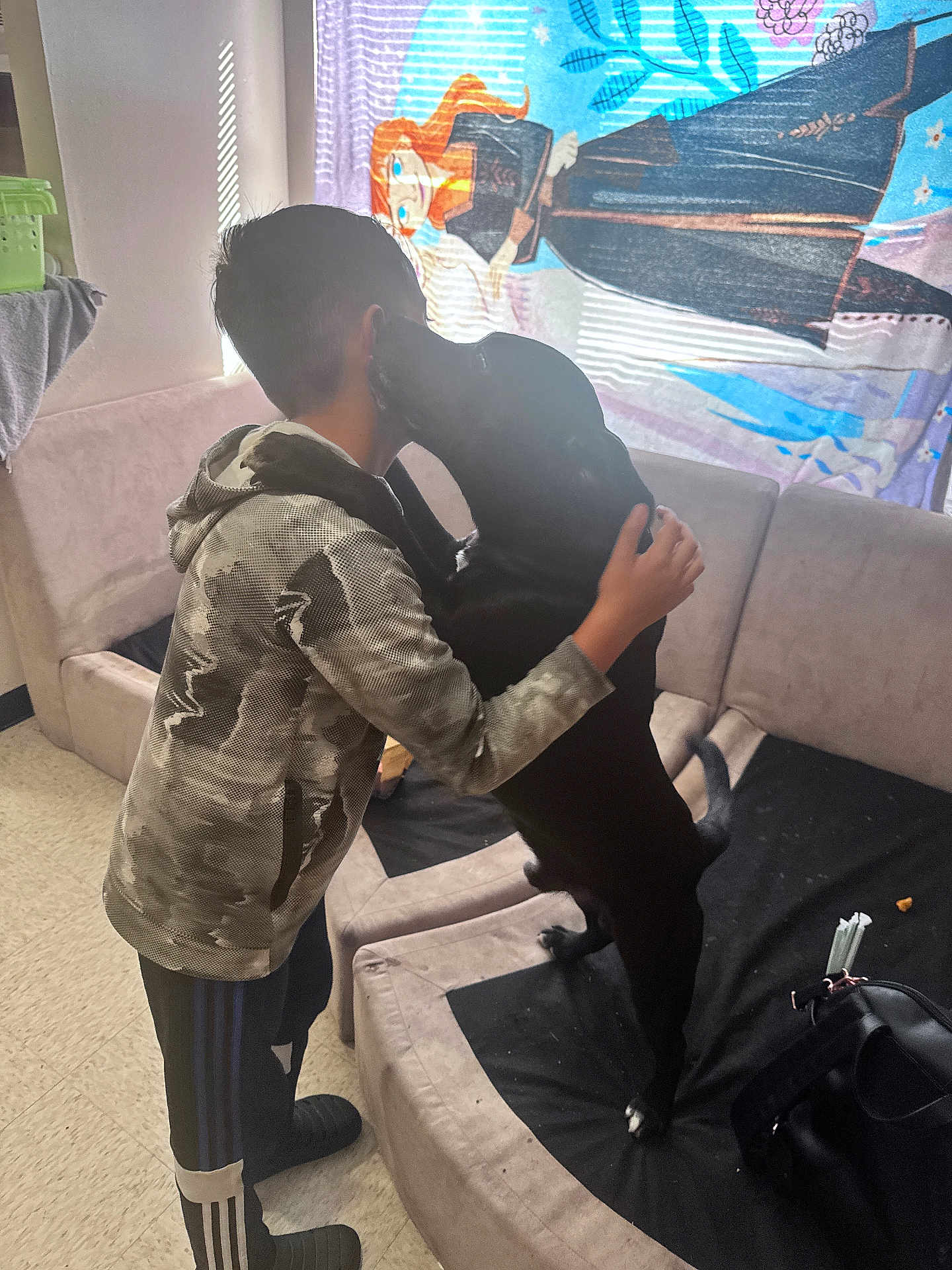 Bluey is registered to the contest to win money with this photo: child, dog, hug, couch, indoor, curtain, cartoon, black_dog, affection, light, window, floor, clothing, sleeves, pants, footwear, back, pet, furniture, home