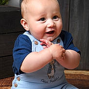 George joined the competition — help win amazing prizes! baby, child, infant, clothing, blue_overalls, peter_rabbit, smiling, happy, hands, face, cute, indoor, wooden_floor, button, short_sleeves, portrait, young_child, sitting, person, light