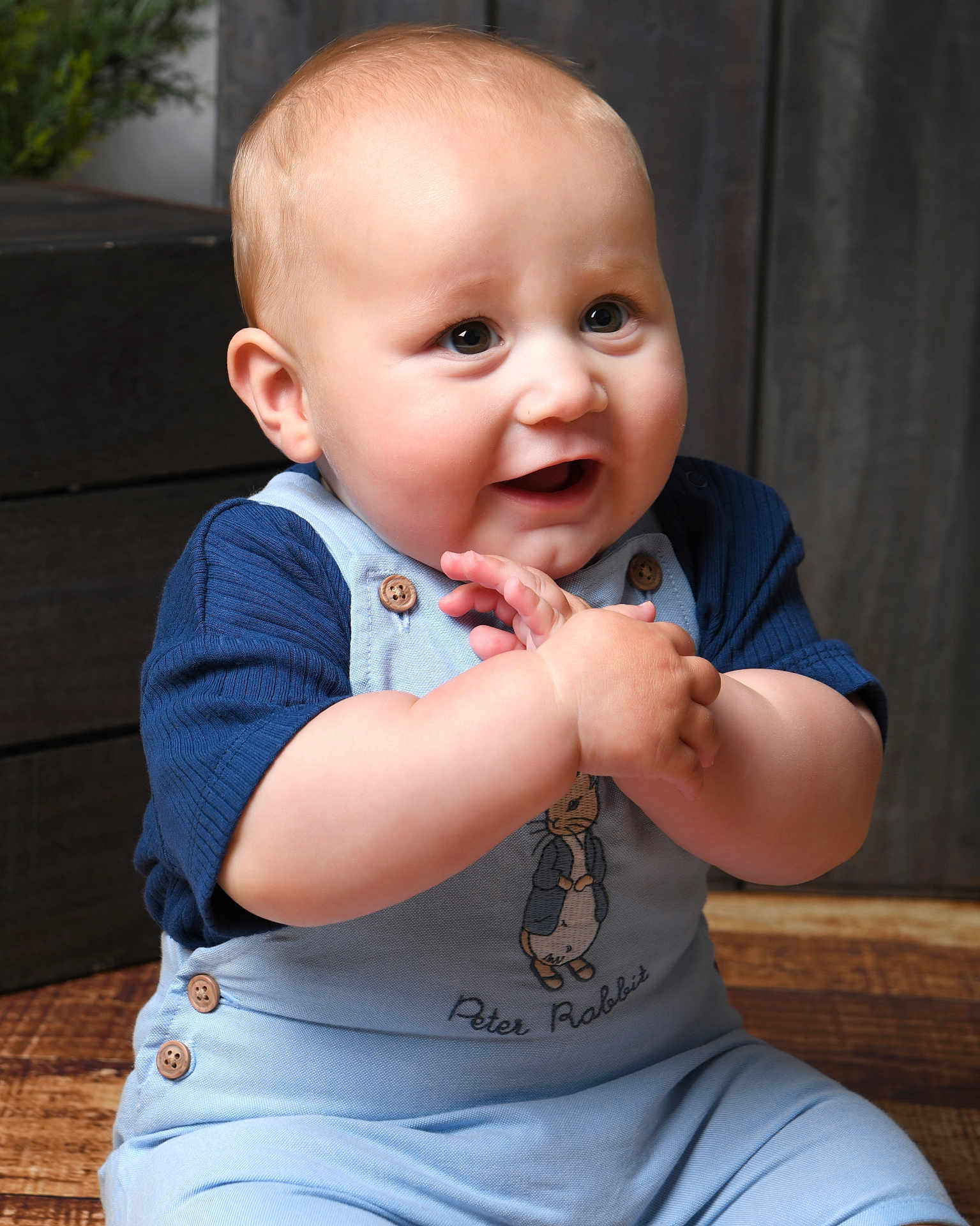 George joined the competition — help win amazing prizes! baby, child, infant, clothing, blue_overalls, peter_rabbit, smiling, happy, hands, face, cute, indoor, wooden_floor, button, short_sleeves, portrait, young_child, sitting, person, light