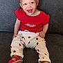 toddler, child, smiling, red_shirt, white_pants, apple_print, red_sandals, polka_dot, sitting, couch, gray_background, happy, cute, indoors, baby, clothing, footwear, portrait, young_child, joyful