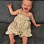 toddler, child, smiling, laughing, dress, hair_clip, barefoot, sitting, couch, grey_fabric, cute, happy, indoors, portrait, baby, one_person, casual, clothing, young_child, natural_light