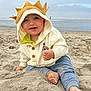 toddler, child, beach, sand, sweater, hood, crown, thumbs_up, happy, smiling, barefoot, ocean, sky, cloudy, outdoor, playing, casual_clothing, cute, portrait, young_child