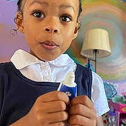 Nayeli Skye joined the competition — help win amazing prizes! child, girl, face, hands, tube, cream, white_shirt, navy_dress, indoor, lamp, colorful_wall, painting, smile_drawing, curly_hair, expression, person, closeup, holding, young, room