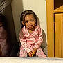 child, toddler, smile, pajamas, pink_clothing, braids, marker, indoor, carpet, cabinet, wood_furniture, wall, happy, sitting, person, cute, playful, beads, young_child, home