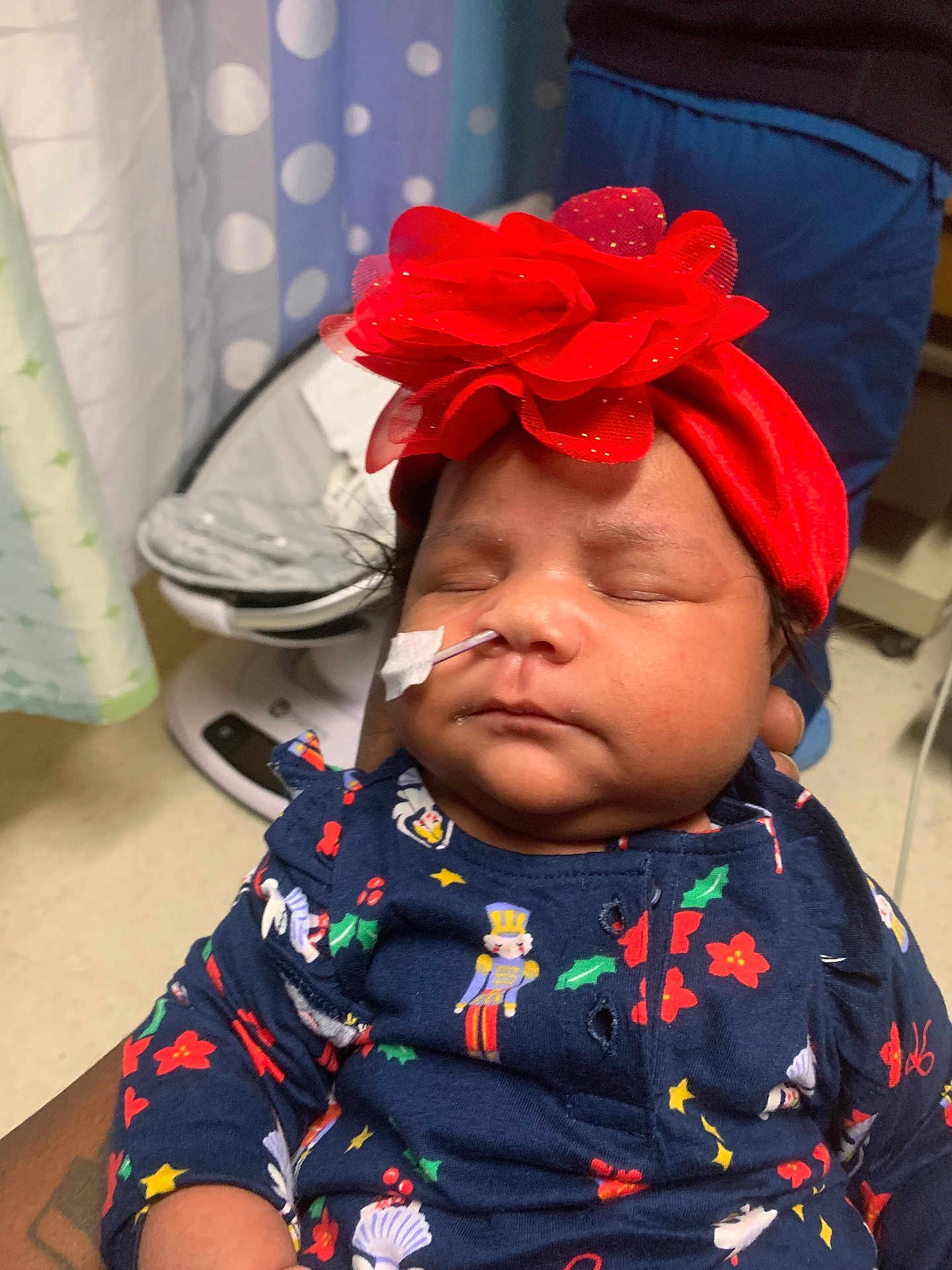 Winter is registered to the contest to win money with this photo: baby, infant, sleeping, headband, flower, medical_tube, tape, navy_blue_clothing, holiday_prints, person_holding, indoor, curtain, baby_rocker, closeup, cute, peaceful, child, newborn, soft_lighting, portrait