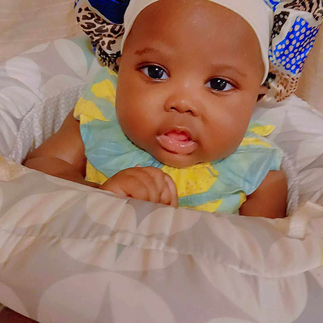 Thiea is registered to the contest to win money with this photo: baby, bonnet, child, clothing, comfort, cushion, cute, expression, face, hand, headwear, indoors, infant, looking, portrait, resting, seated, skin, soft, young