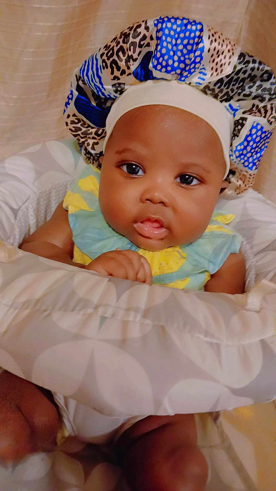 Thiea is registered to the contest to win money with this photo: baby, infant, child, bonnet, cushion, seated, indoors, clothing, face, portrait, cute, looking, hand, skin, headwear, young, expression, soft, comfort, resting