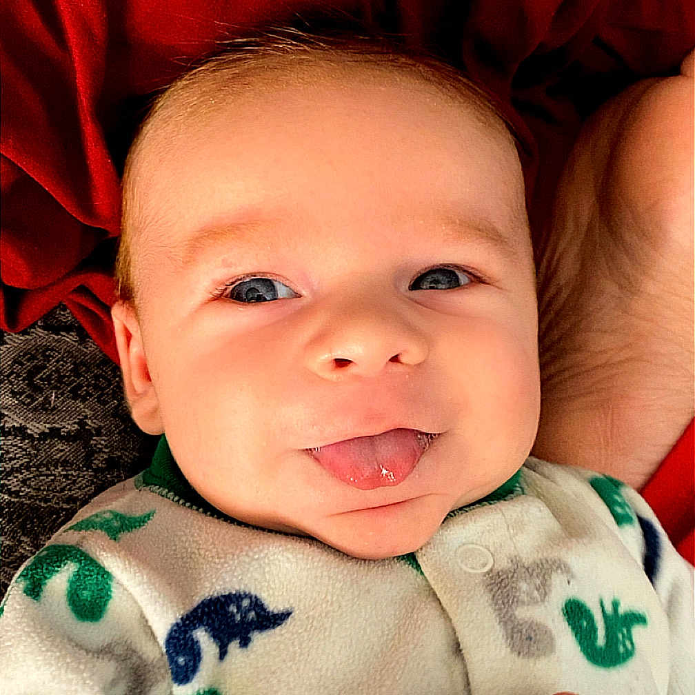 Axel Ryan is registered to the contest to win money with this photo: baby, child, clothing, cozy, cute, dinosaur_pattern, expression, eyes, face, hand, infant, lying_down, onesie, person, playful, portrait, red_background, skin, smiling, tongue_out