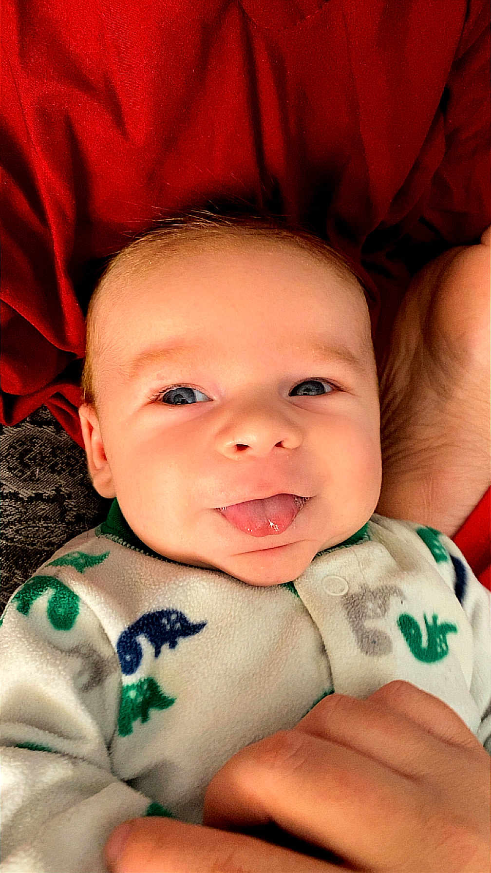 Axel Ryan is registered to the contest to win money with this photo: baby, infant, tongue_out, onesie, dinosaur_pattern, hand, red_background, face, smiling, cute, child, skin, eyes, person, cozy, clothing, expression, portrait, lying_down, playful