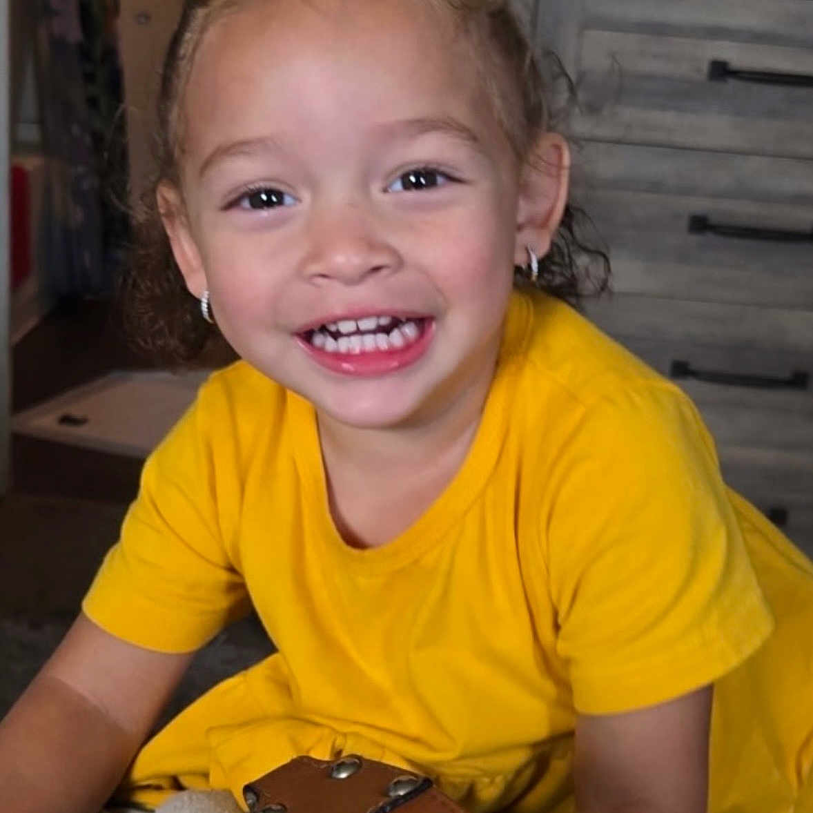 Macy is registered to the contest to win money with this photo: child, toddler, smiling, yellow_shirt, yellow_bows, indoor, portrait, happy, face, curly_hair, earrings, drawer, furniture, toy, bag, playing, person, room, home, cute