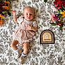Scout joined the competition — help win amazing prizes! baby, infant, child, plaid_dress, shoes, ruffled_socks, blanket, patterned_blanket, flowers, floral_arrangement, wooden_plaque, milestone_marker, cute, laying_down, indoors, portrait, adorable, young_child, soft_lighting, celebration