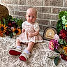 Scout joined the competition — help win amazing prizes! baby, infant, child, flower, sunflower, dress, shoes, socks, brick_wall, tablecloth, floral_arrangement, greenery, wooden_sign, milestone, portrait, indoor, cute, sitting, decor, colorful