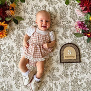 Scout is registered to the contest to win money with this photo: baby, infant, smiling, gingham, dress, shoes, socks, blanket, pattern, flowers, floral_arrangement, wooden_sign, months, cute, happy, lying_down, portrait, indoor, child, person